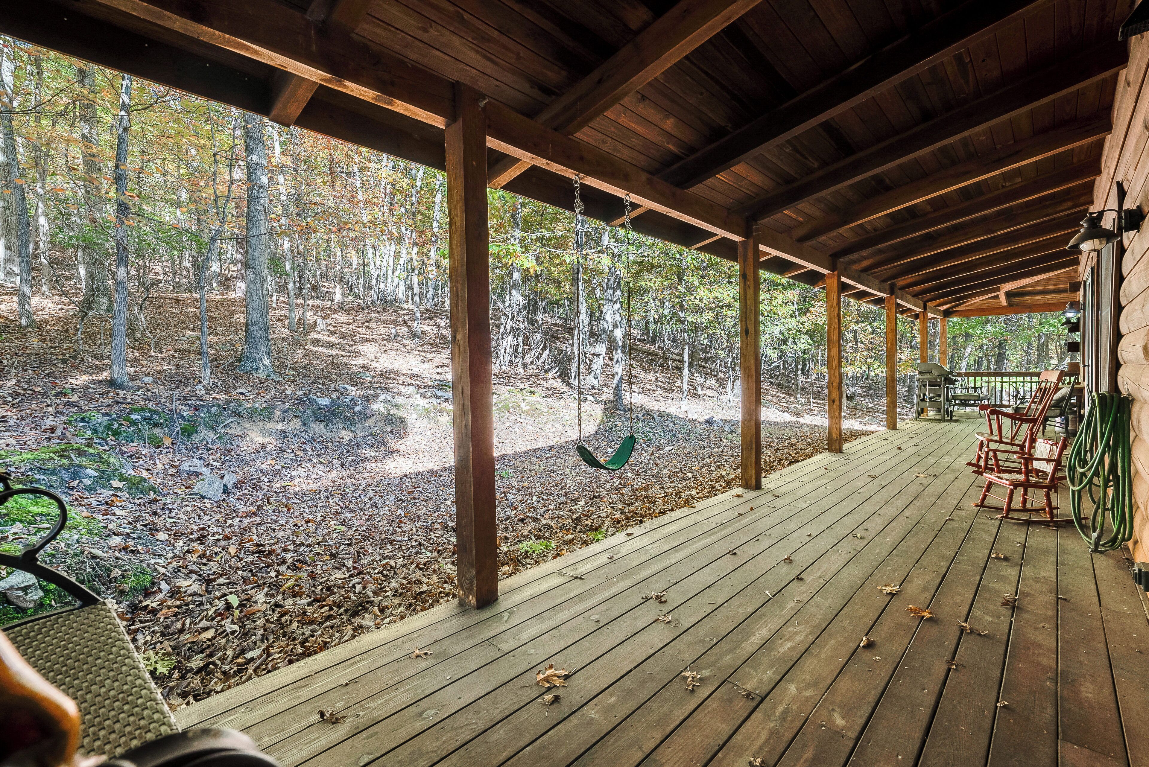 A covered porch with a wooden deck, a swing, and a view of a forested area.