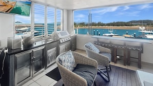 Een balkon met uitzicht op de jachthaven, een keukenhoek met een kookplaat en kasten, rieten stoelen en een tafel.