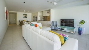 Een moderne woonkamer met een witte bank, een televisie en een eethoek op de achtergrond.