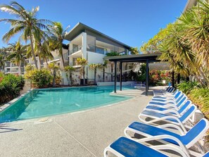 A swimming pool with lounge chairs, palm trees, and a building with balconies.