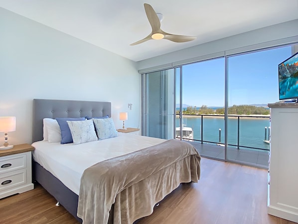 A bedroom with a large bed, a ceiling fan, a TV, and a view of the water.