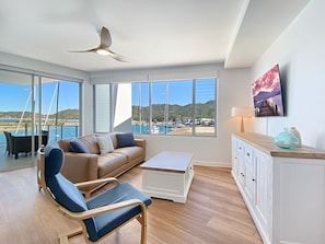 A spacious living area with a large window, a ceiling fan, a sofa, a coffee table, and a TV mounted on the wall.