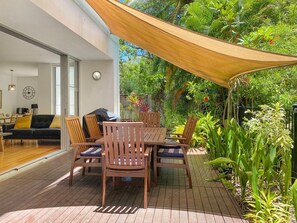A patio with a wooden table and chairs, surrounded by lush greenery and a large shade sail. - Maggies Beachfront Apt. 9 (Horseshoe Bay)
