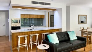 A modern kitchen with a central island, wooden stools, a black leather sofa with teal cushions, and a round table with a remote control.