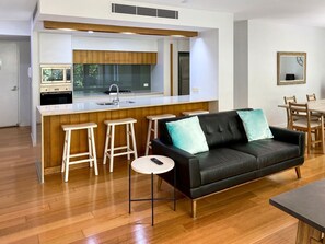 A modern kitchen with a central island, wooden stools, a black leather sofa with teal cushions, and a round table with a remote control.