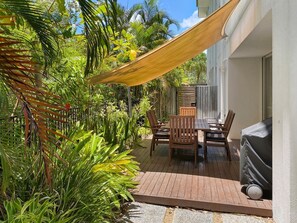 A patio with a table and chairs, surrounded by lush greenery and a wooden deck.