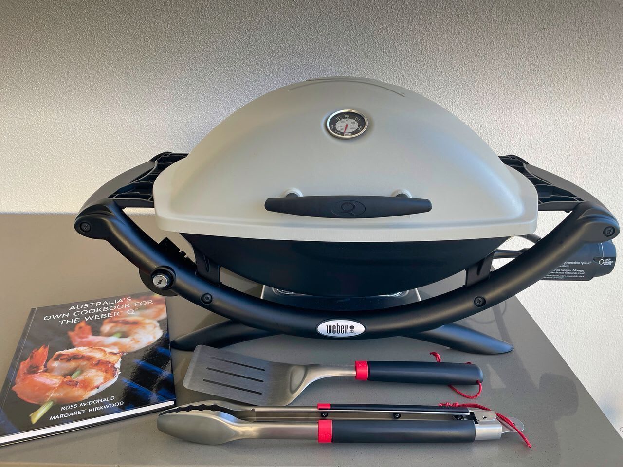 Ein Weber-Grill mit Kochbuch, Pfanne und Grillzange.