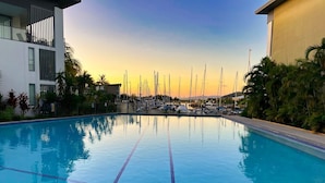 A swimming pool with clear blue water, surrounded by a fence, and a view of boats in the background.