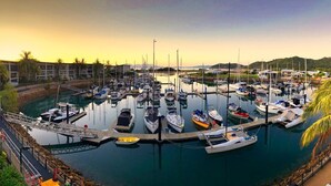 A marina with multiple boats docked, surrounded by a hotel with a pool and palm trees.