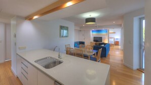 A modern kitchen with a central island, a dining area with wooden chairs, a sofa, and a TV in the background. - Maggies Beachfront Apt. 6 (Horseshoe Bay)