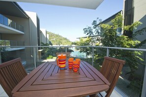 A balcony with a table and chairs, a view of buildings and greenery, and a pool in the background.