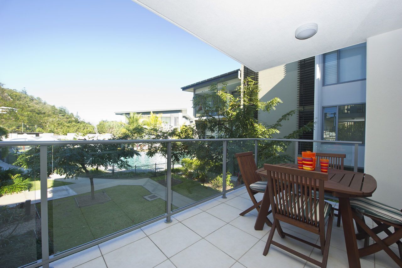 A balcony with a table and chairs, a glass railing, and a view of a pool and greenery.