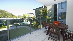 A balcony with a table and chairs, a glass railing, and a view of a pool and greenery. - Blue on Blue Apartment 921 (Nelly Bay)