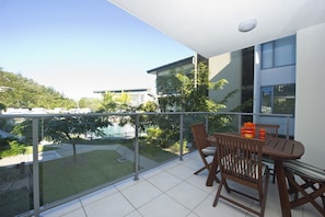 Ein Balkon mit Tisch, Stühlen, einer Glastrennwand und Blick auf ein Schwimmbecken und Grünflächen.