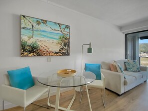 Ein modernes Wohnzimmer mit einem Glastisch, wei ßen Sofas und einem Wandbild mit Strandmotiv.