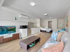 A modern living room with a flat-screen TV, a wooden coffee table, a grey sofa with patterned cushions, a kitchen area with white cabinets, and a dining area with a table and chairs. - Blue on Blue Superior Studio Room 1241 (Nelly Bay)