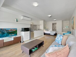 Ein modernes Wohnzimmer mit Flachbildfernseher, einem Holztisch, einem grauen Sofa mit gemusterten Kissen, einer Küchenzeile mit weißen Schränken sowie einem Essbereich mit Tisch und Stühlen.