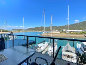 A marina with boats docked, a table and chairs on a balcony, and a view of the water and hills. - Blue on Blue Superior Studio Room 1241 (Nelly Bay)