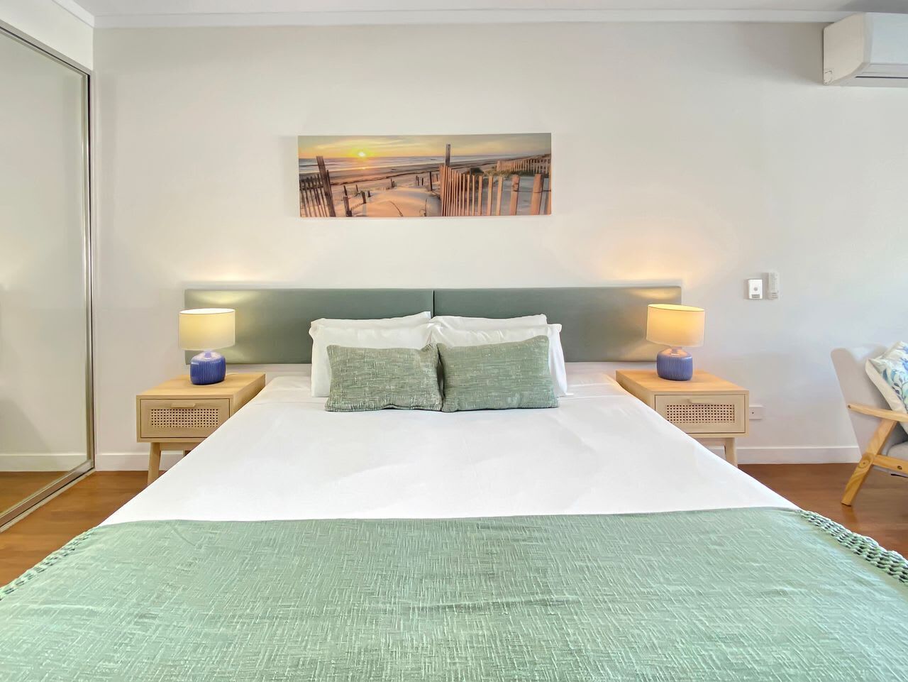 A bedroom with a bed, two bedside tables with lamps, a headboard, a wall-mounted artwork, and a chair.