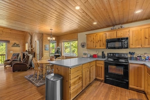 A cabin interior with a wooden ceiling, a kitchen with wooden cabinets, a dining area with a table and chairs, and a fireplace.