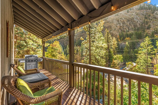 A covered outdoor area with a grill, wicker furniture, and a view of a forested mountain.