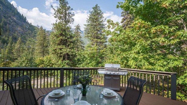 An outdoor dining area with a table set for four, surrounded by a deck with a railing, overlooking a forested mountain area.