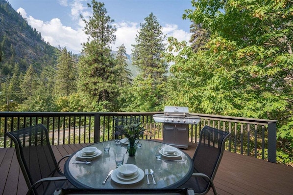 Un área de comedor al aire libre con una mesa preparada para cuatro, rodeada por una terraza con barandilla y vistas a una zona montañosa boscosa.