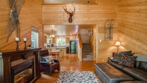 A log cabin interior with a fireplace, leather seating, a kitchen area, and a staircase leading to another level.
