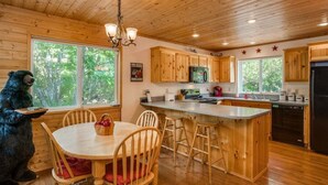 A wooden cabin interior with a dining table, chairs, a kitchen area with wooden cabinets, a refrigerator, and a bear figurine.