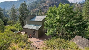 A two-story cabin with a metal roof, surrounded by dense forest and mountains.