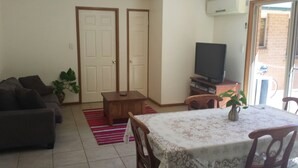 Una sala de estar con un sofá, una mesa de centro, un televisor y una mesa de comedor con sillas. Hay dos puertas y una ventana con vistas a un edificio de ladrillo.