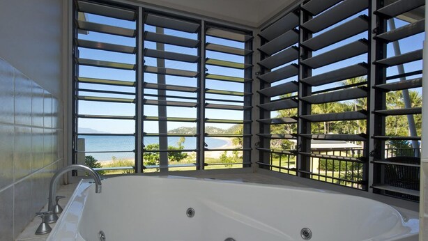 Una bañera amplia con vista al mar a través de una ventana con persianas horizontales.