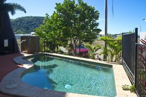 A swimming pool with a curved edge, surrounded by a fence and trees, with a view of a hilly landscape in the background.