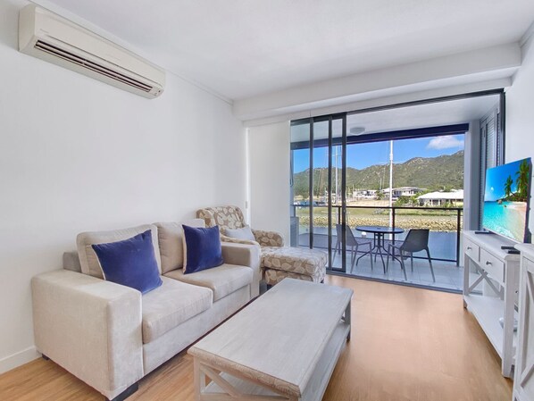 A modern living room with a sofa, coffee table, and a TV. Large windows offer a view of the sea and buildings.