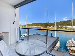 A balcony with a glass table and chairs, overlooking a marina with boats and a hilly landscape.