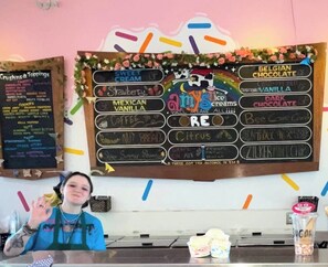 Eine Kreidetafel zeigt verschiedene Eissorten, dahinter steht eine Person an der Theke.