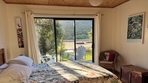 Une chambre avec un grand lit, une chaise en osier, une petite armoire en bois et une vue sur un jardin, accessible par une porte coulissante.