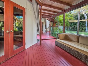 A covered outdoor area with a red wooden floor, a cushioned bench, and a glass door leading to an interior space. - Serene Place (Nelly Bay)