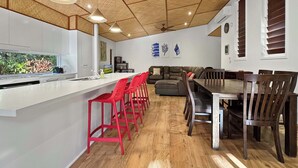A modern kitchen with a white countertop, red bar stools, a dining table with wooden chairs, and a ceiling fan. - Serene Place (Nelly Bay)