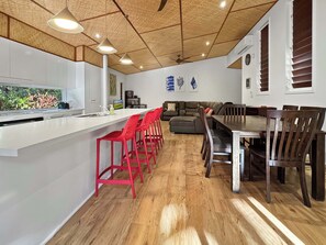 A modern kitchen with a white countertop, red bar stools, a dining table with wooden chairs, and a ceiling fan. - Serene Place (Nelly Bay)