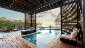 A pool area with wooden decking, lounge chairs, and a view of the ocean at sunset.