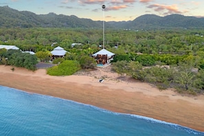 An aerial view of a coastal property with a beach, a large house with a distinctive roof, and surrounding greenery.