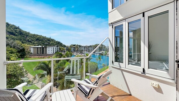 Ein Balkon mit zwei weißen Liegestühlen, einer Glasreling und Blick auf einen Yachthafen und Grünflächen.