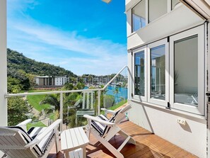 Ein Balkon mit zwei weißen Liegestühlen, einer Glasreling und Blick auf einen Yachthafen und Grünflächen.