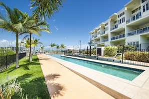 A swimming pool area with a clear sky, palm trees, and a multi-story building with balconies. - Beachside Apartment 14 (Nelly Bay)