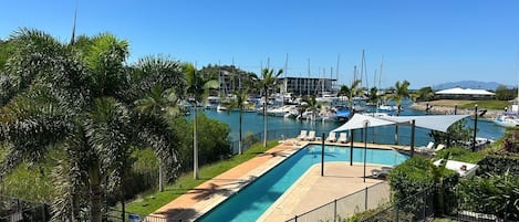 Una piscina con sillones de descanso, rodeada de palmeras y un puerto deportivo con barcos.