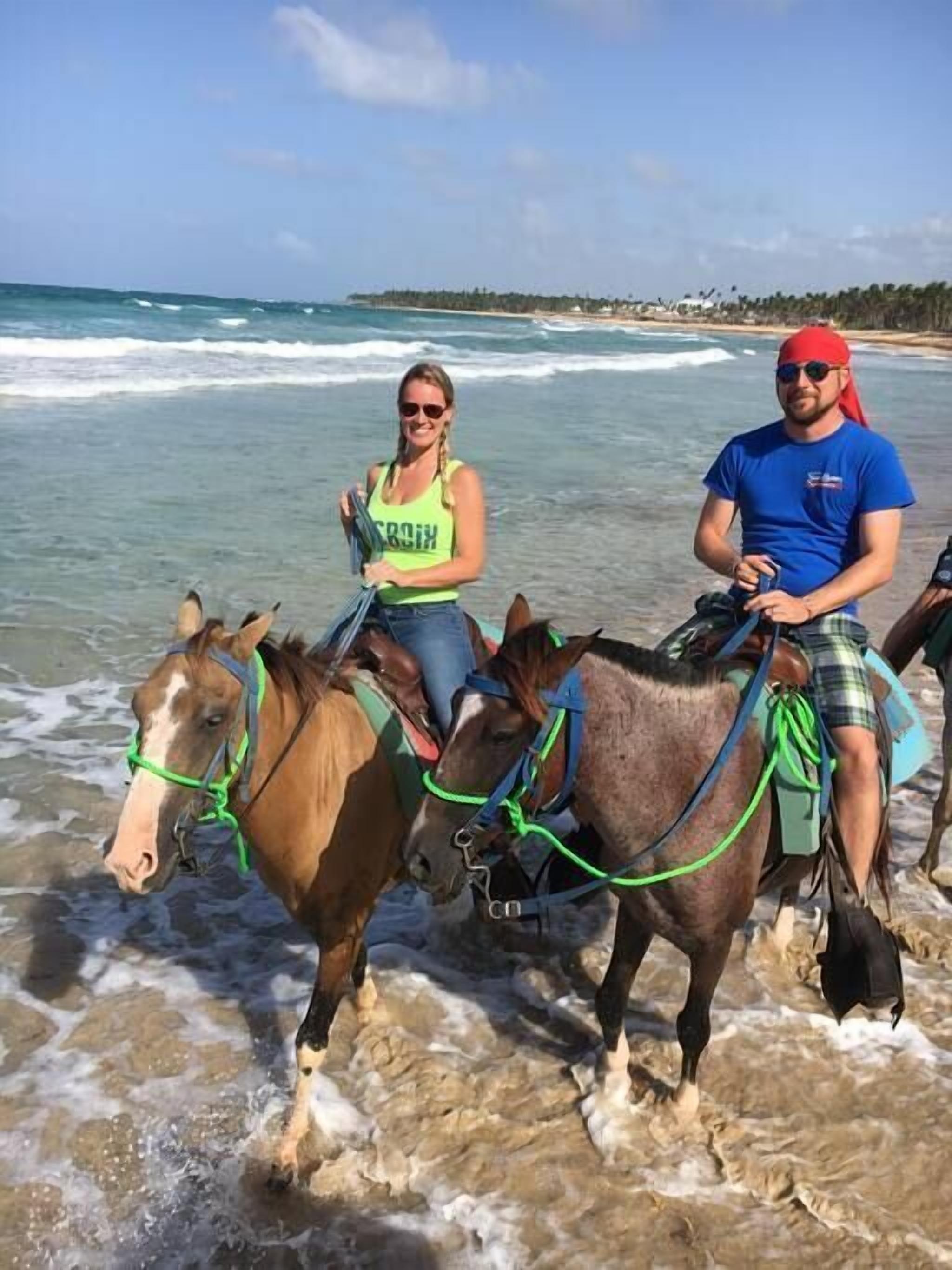 Dos personas montando a caballo en una playa con olas del océano al fondo.
