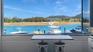 A room with a large window overlooking a marina with boats, a sandy beach, and a hilly landscape.