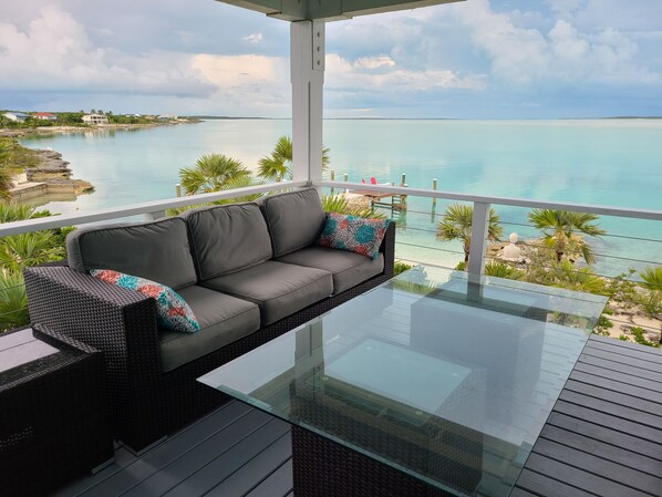 A balcony with a glass-top table and a grey sofa with colorful cushions, overlooking a turquoise sea and a clear sky.