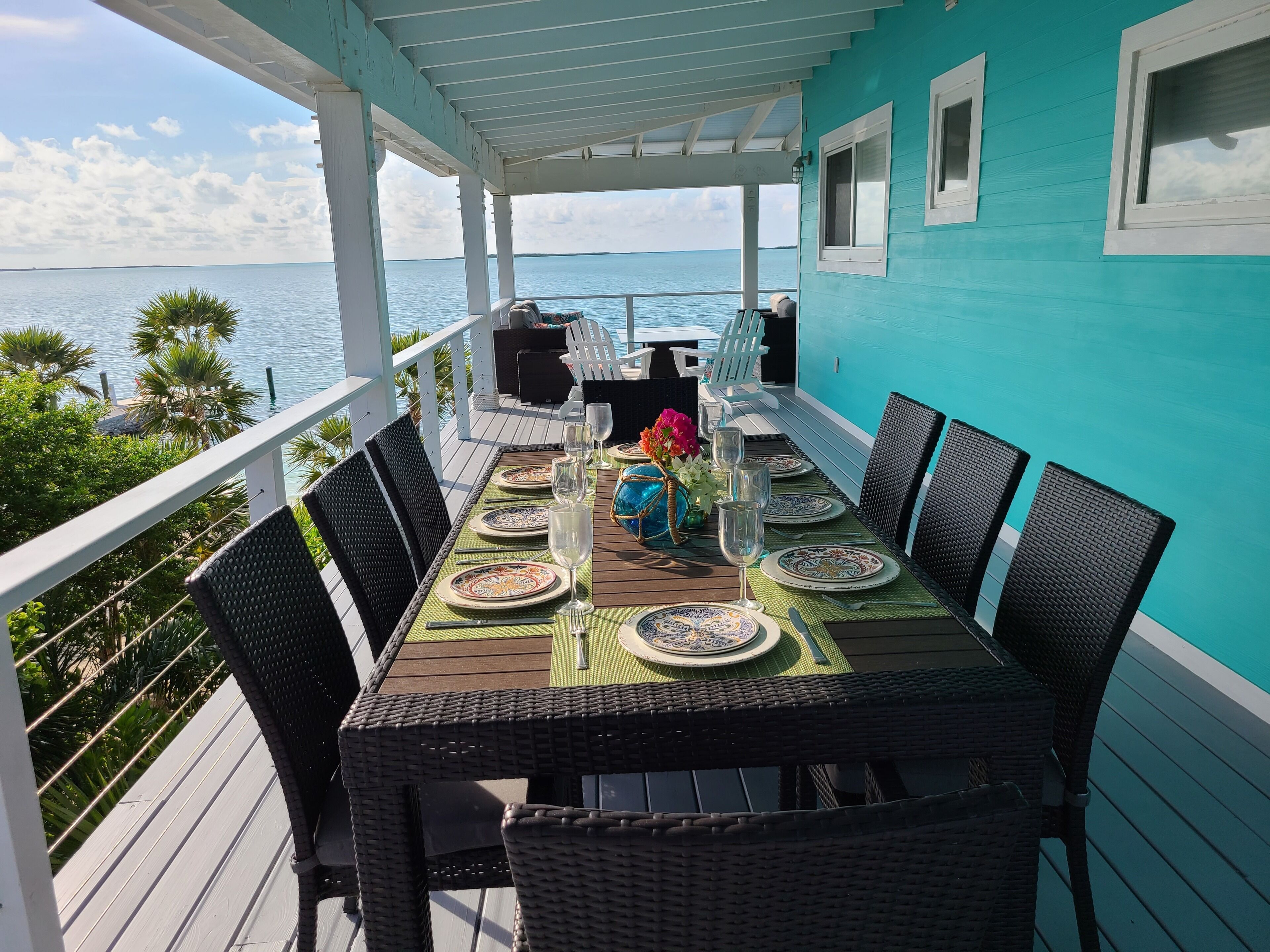 A covered outdoor dining area with a view of the sea, featuring a table set for four with chairs, a decorative vase, and a clear sky.